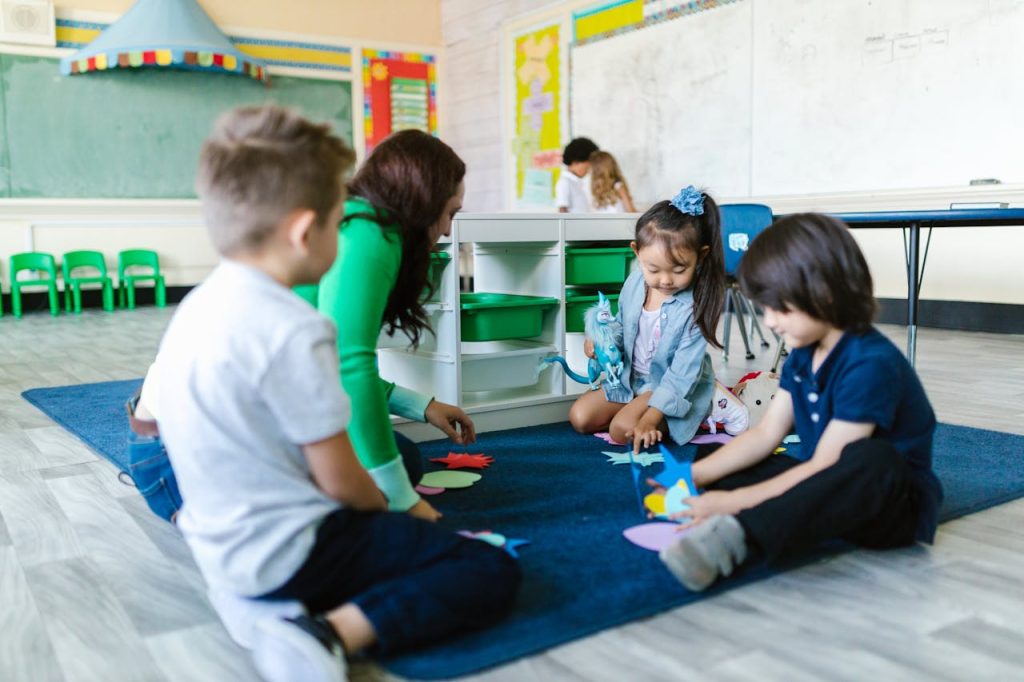 children-sitting-on-a-carpet-8363152 Kids and teacher in a kindergarten classroom engaging in creative play on the carpet.