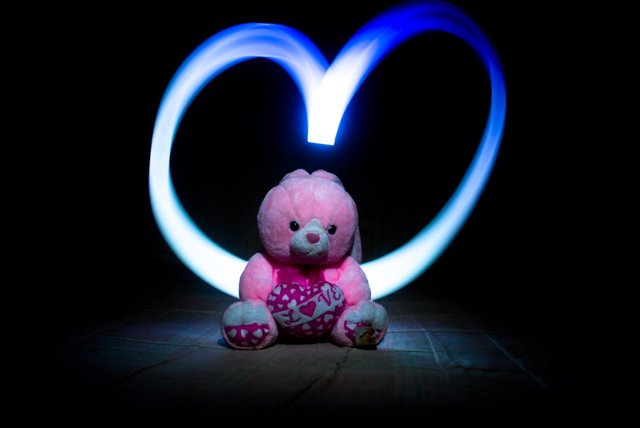 Adorable pink teddy bear with heart-shaped light painting in a dark setting, symbolizing love.